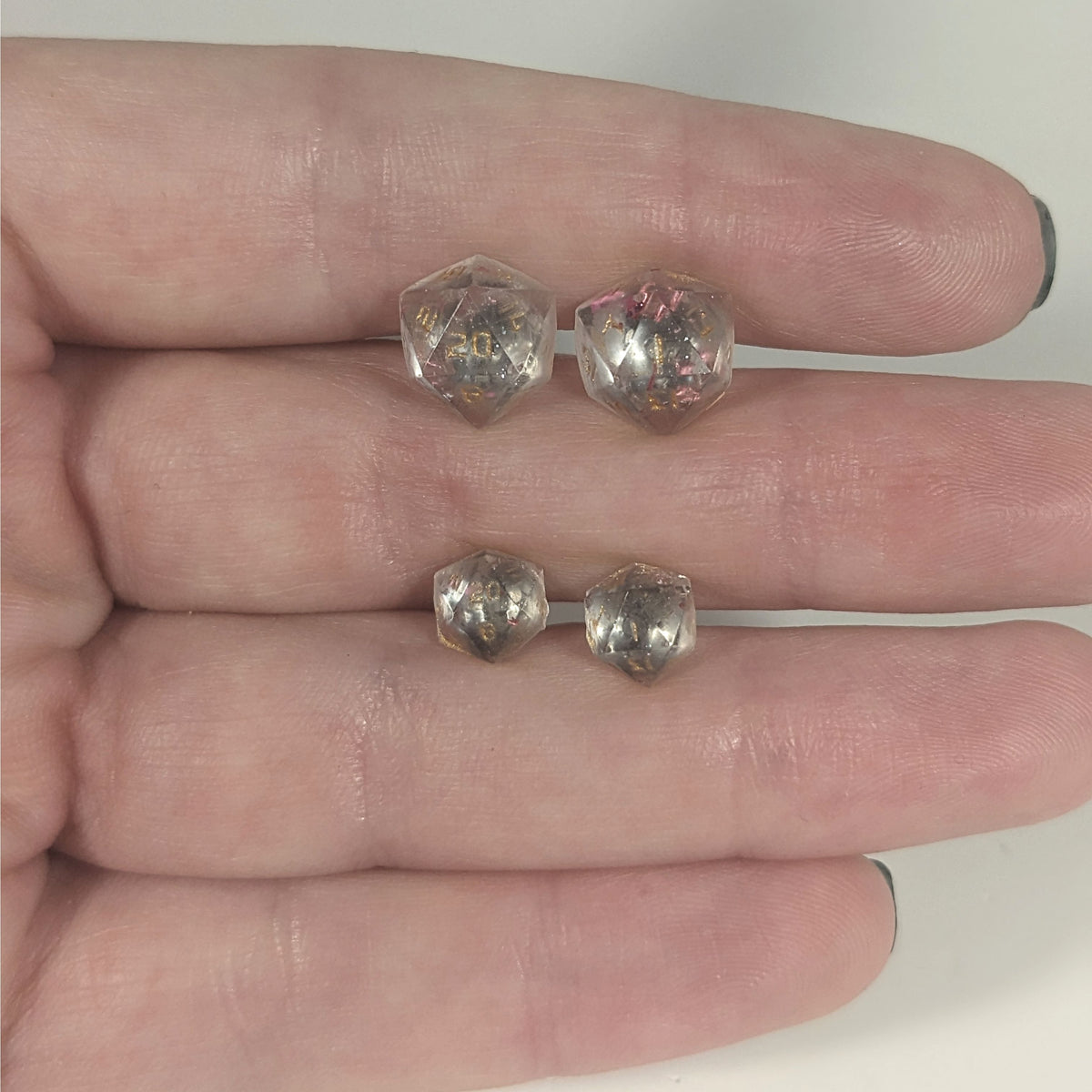 Two pairs of handmade d20 earrings with titanium hardware that are glittery clear with pink flakies with gold numbers held between the fingers of a flat hand. The larger pair is between the pointer and middle finger, the smaller between the middle and ring finger. The earrings on the left of each pair centers the 20 face and the earrings on the right of each pair centers the 1 face.