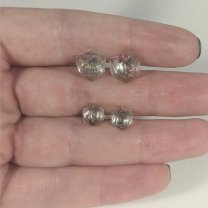 Two pairs of handmade d20 earrings with titanium hardware that are glittery clear with pink flakies with gold numbers held between the fingers of a flat hand. The larger pair is between the pointer and middle finger, the smaller between the middle and ring finger. The earrings on the left of each pair centers the 20 face and the earrings on the right of each pair centers the 1 face.