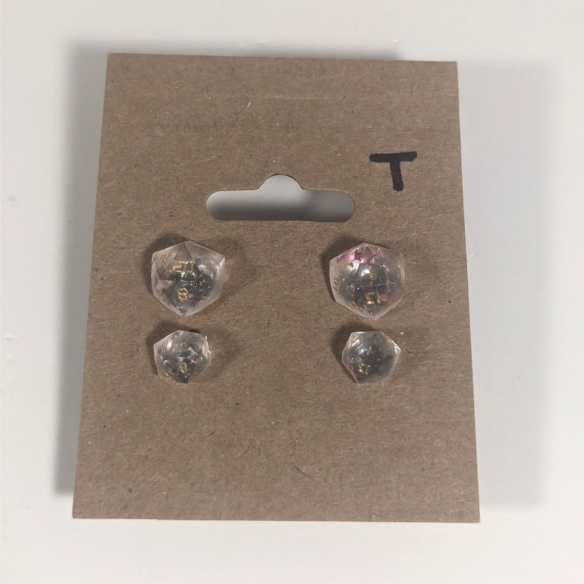 Two pairs of handmade d20 earrings with titanium hardware that are glittery clear with pink flakies with gold numbers on a kraft brown earring card with a T in the top right corner. The earrings on the left of each pair centers the 20 face and the earrings on the right of each pair centers the 1 face.
