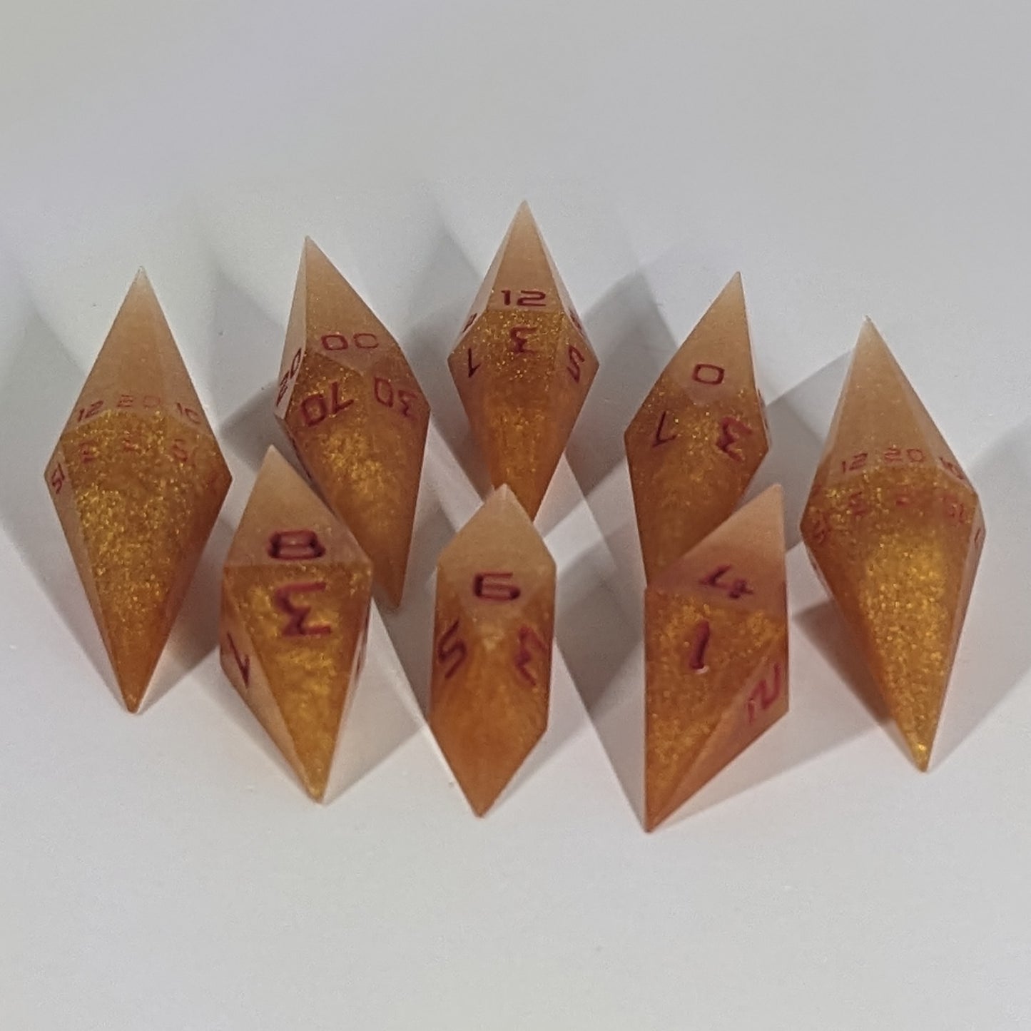  A complete set of resin dice featuring 2 d20s for advantage rolls. These shimmering gold and red gold swirls and red numbering slender handmade diamond dice are clustered in a tight circle on a white background, highlighting the quality of the artisan dice craftsmanship and the unique pointed shape.