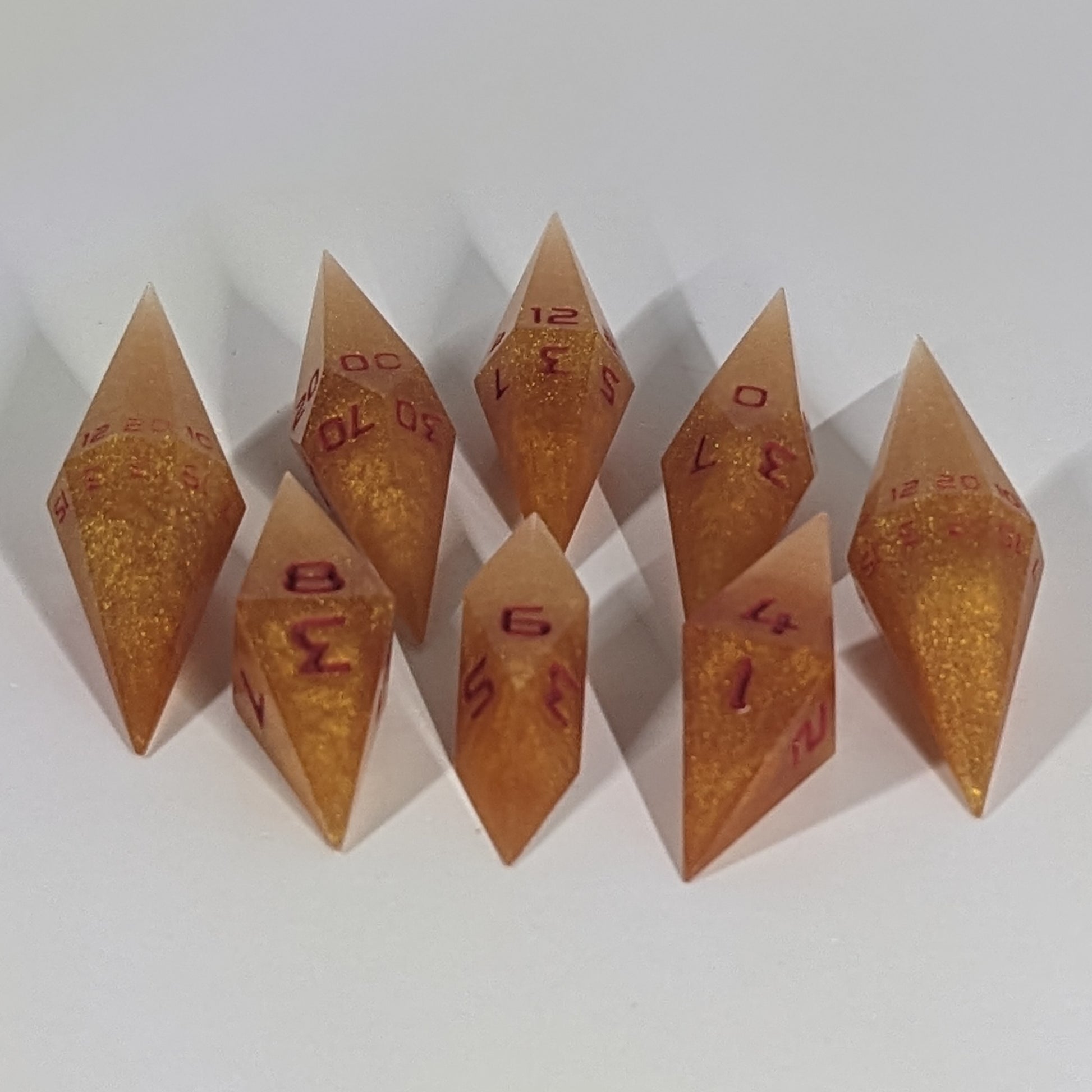  A complete set of resin dice featuring 2 d20s for advantage rolls. These shimmering gold and red gold swirls and red numbering slender handmade diamond dice are clustered in a tight circle on a white background, highlighting the quality of the artisan dice craftsmanship and the unique pointed shape.