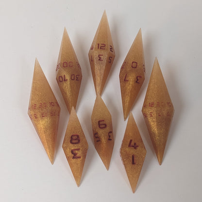 A complete set of resin dice featuring 2 d20s for advantage rolls. These shimmering gold and red gold swirls and red numbering slender handmade diamond dice are clustered in a tight circle on a white background, highlighting the quality of the artisan dice craftsmanship and the unique pointed shape.