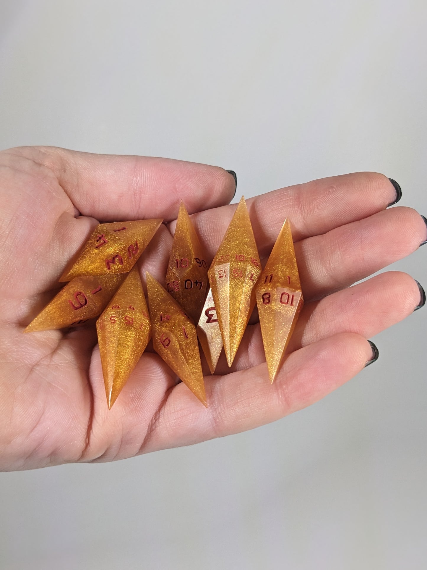 Close-up of shimmering gold and red gold swirls and red numbering  artisan dice resting in a palm on a seamless white backdrop. The handmade dice feature a distinct sharp diamond shape, with 2 d20s visible among the high-quality resin dice.