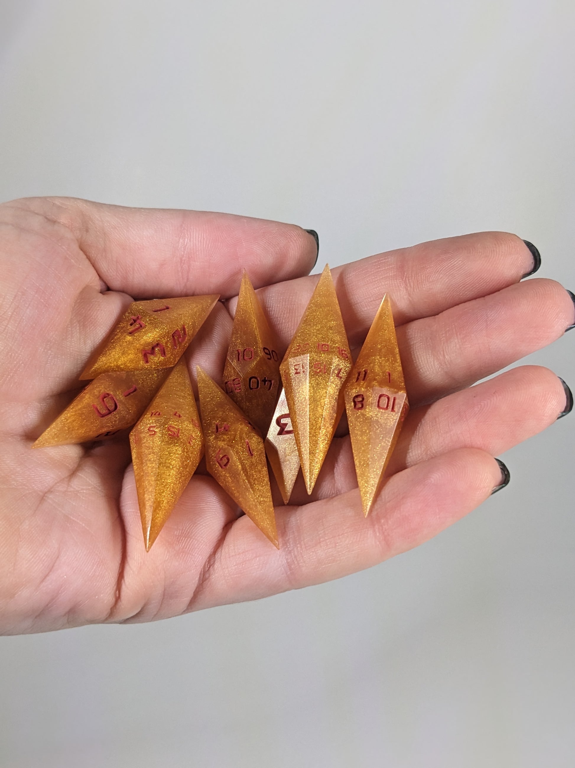 Close-up of shimmering gold and red gold swirls and red numbering  artisan dice resting in a palm on a seamless white backdrop. The handmade dice feature a distinct sharp diamond shape, with 2 d20s visible among the high-quality resin dice.