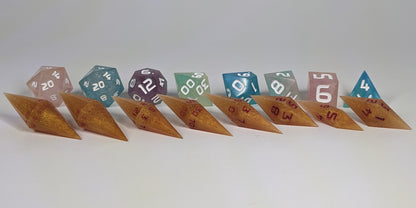 A size comparison of shimmering gold and red gold swirls and red numbering long pointed diamond handmade dice with standard shapes placed behind for scale. The photo emphasizes the length of the diamond dice being much greater than a standard set as well as the diamond set being much more slender. This artisan dice set features 2 d20s and rests on a seamless white background to highlight the elongated profile of the resin dice.