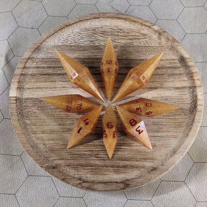 A set of shimmering gold and red gold swirls and red numbering handmade dice arranged in a wooden circle organizer. The resin dice feature a unique pointed diamond shape with the tips facing outward, highlighting the sharp edges of the two d20 and other artisan dice in the collection. 