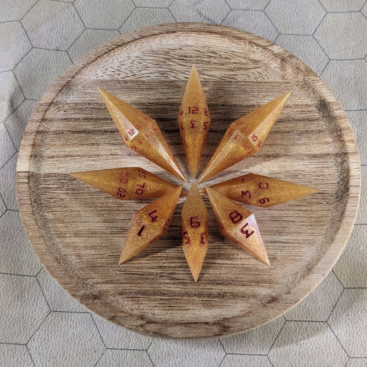 A set of shimmering gold and red gold swirls and red numbering handmade dice arranged in a wooden circle organizer. The resin dice feature a unique pointed diamond shape with the tips facing outward, highlighting the sharp edges of the two d20 and other artisan dice in the collection. 