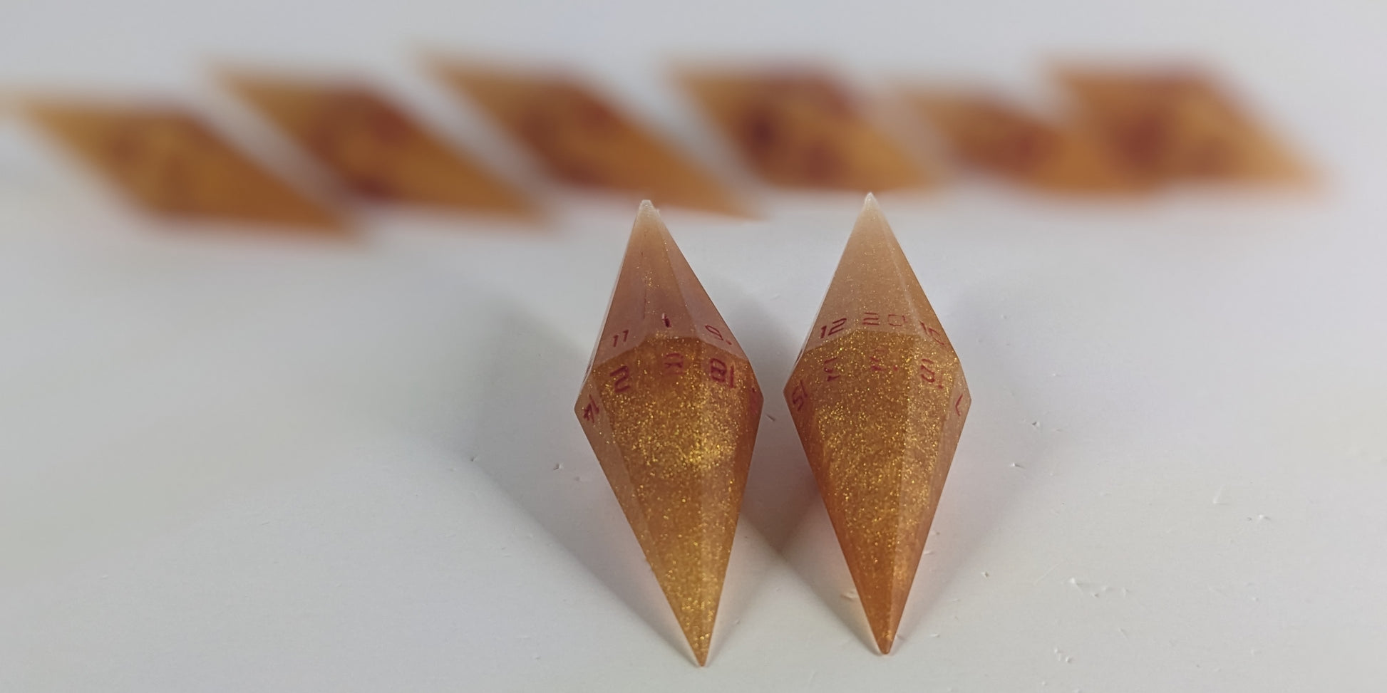 A stylized macro shot focusing heavily on 2 d20s from a shimmering gold and red gold swirls and red numbering handmade dice set in the foreground. The sharp diamond edges of these front resin dice are crisp, while the rest of the matching artisan dice collection forms a softly blurred background, creating depth.