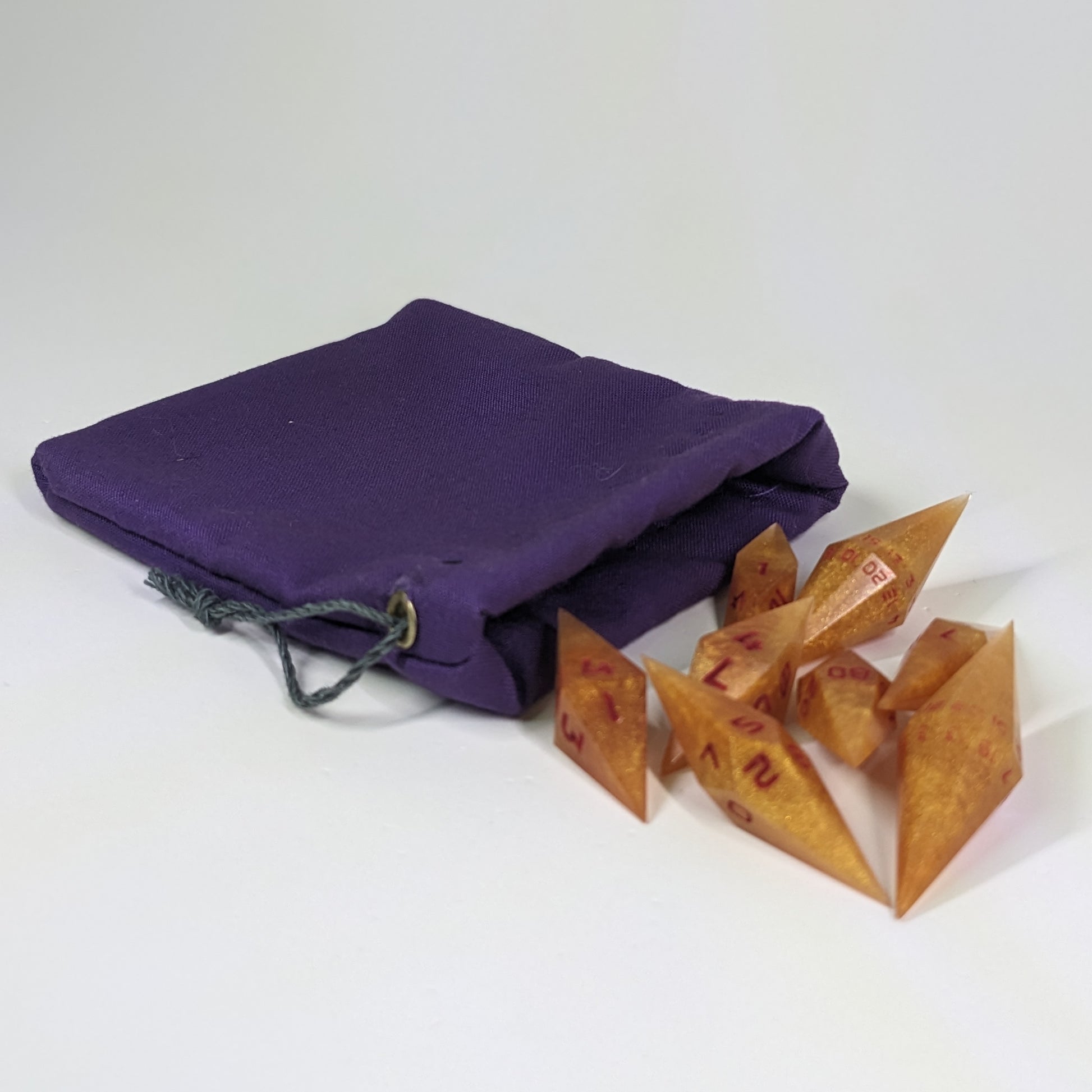 Shimmering gold and red gold swirls and red numbering diamond handmade dice spilling out of a purple drawstring bag on a white background. The set features 2 d20s and matching resin dice, showcasing the unique shape of these artisan dice.