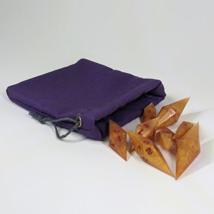Shimmering gold and red gold swirls and red numbering diamond handmade dice spilling out of a purple drawstring bag on a white background. The set features 2 d20s and matching resin dice, showcasing the unique shape of these artisan dice.