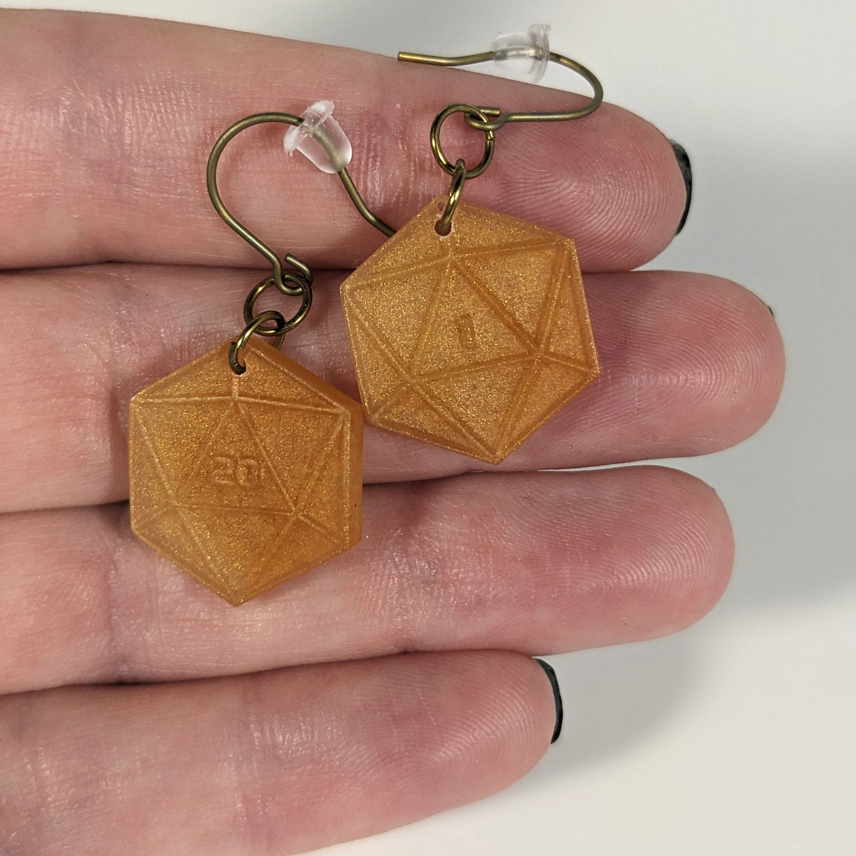 A pair of sparkly gold dangly, flat d20 earrings with gold color titanium and niobium hardware with bell shaped rubber backs held on on a hand on a white background. The earring on the left has a 20 on it and the earring on the right has a 1 on it, both in a sci-fi style font.