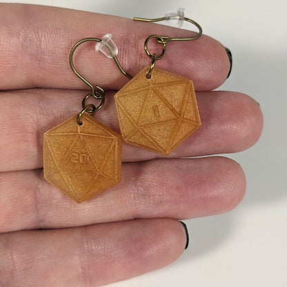 A pair of sparkly gold dangly, flat d20 earrings with gold color titanium and niobium hardware with bell shaped rubber backs held on on a hand on a white background. The earring on the left has a 20 on it and the earring on the right has a 1 on it, both in a sci-fi style font.