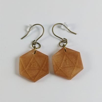 A pair of sparkly gold dangly, flat d20 earrings with gold color titanium and niobium hardware and bell shaped rubber backs on a white background. The earring on the left has a 20 on it and the earring on the right has a 1 on it, both in a fantasy style font.