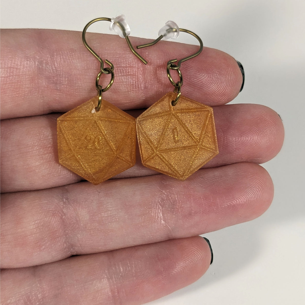 A pair of sparkly gold dangly, flat d20 earrings with gold color titanium and niobium hardware with bell shaped rubber backs held on on a hand on a white background. The earring on the left has a 20 on it and the earring on the right has a 1 on it, both in a fantasy style font.