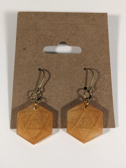 A pair of sparkly gold dangly, flat d20 earrings with gold color titanium and niobium hardware on a kraft earring card on a white background. The earring on the left has a 20 on it and the earring on the right has a 1 on it, both in a fantasy style font.
