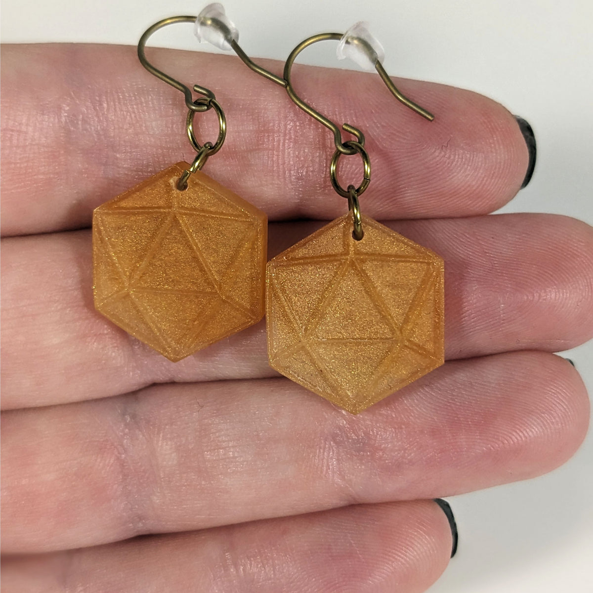 A pair of sparkly gold dangly, flat d20 earrings with gold color titanium and niobium hardware with bell shaped rubber backs held on on a hand on a white background.