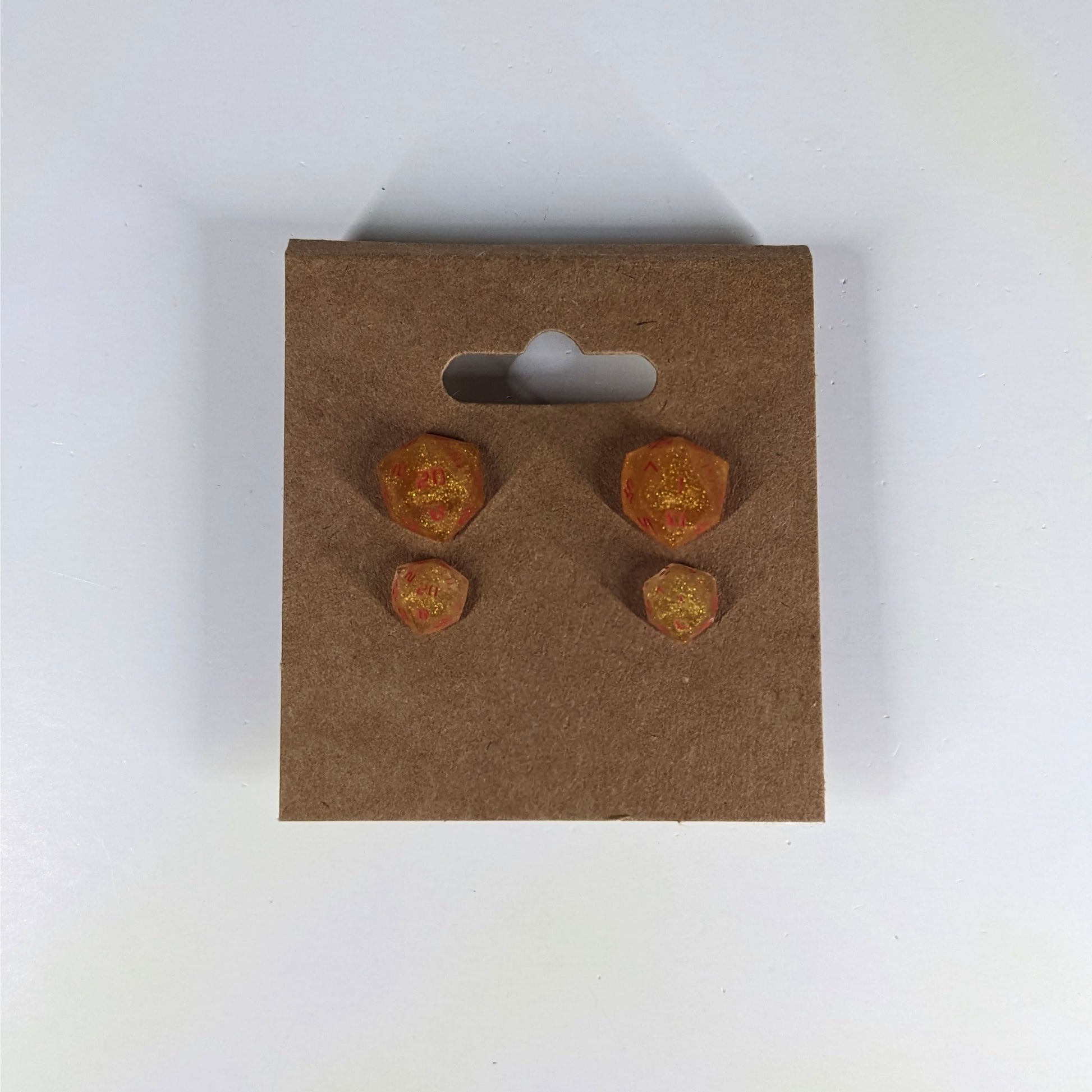 Two pairs of handmade d20 earrings that are shimmering gold with red numbers on a kraft brown earring card. The earrings on the left of each pair centers the 20 face and the earrings on the right of each pair centers the 1 face.