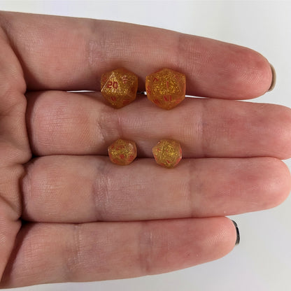 Two pairs of handmade d20 earrings that are shimmering gold with red numbers held between the fingers of a flat hand. The larger pair is between the pointer and middle finger, the smaller between the middle and ring finger. The earrings on the left of each pair centers the 20 face and the earrings on the right of each pair centers the 1 face.