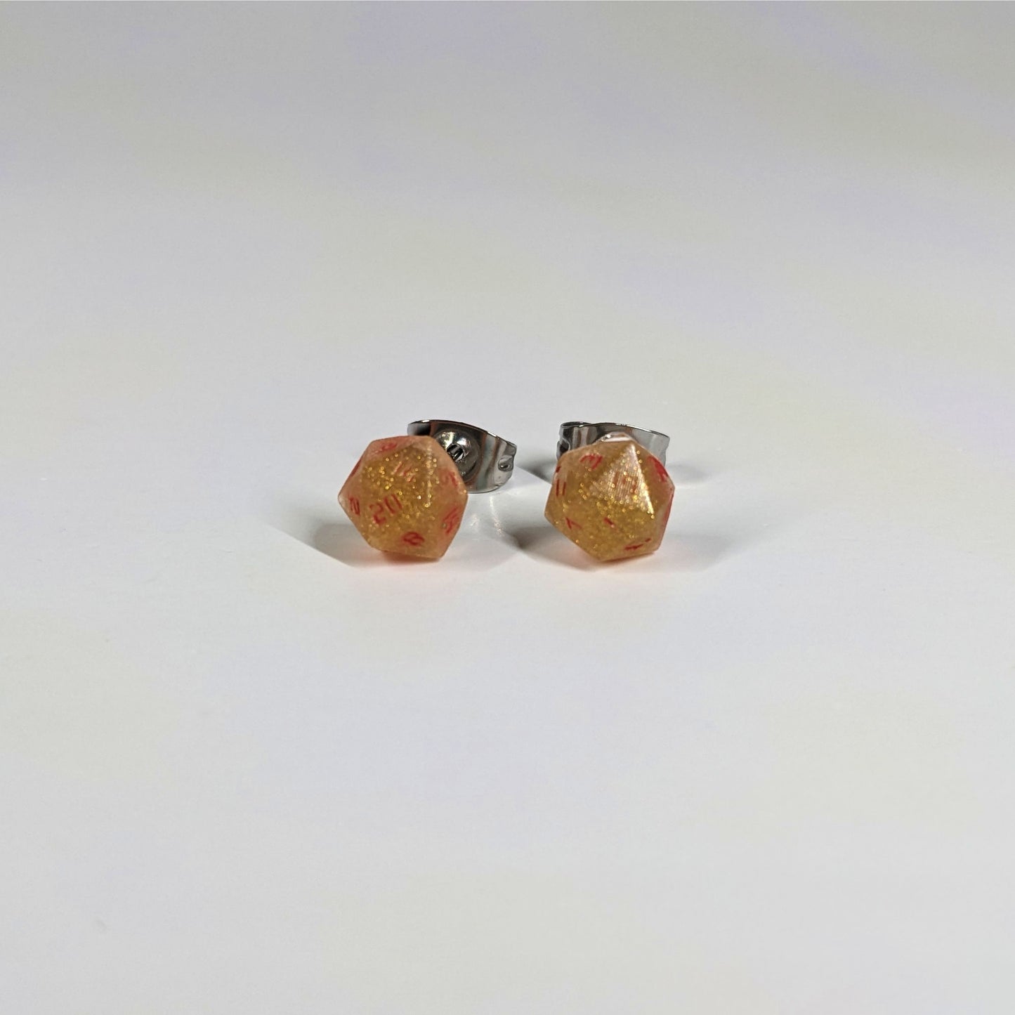 A single pair of the smaller size handmade d20 earrings that are shimmering gold with red numbers on a white background. The earring on the left centers the 20 face and the earring on the right centers the 1 face.