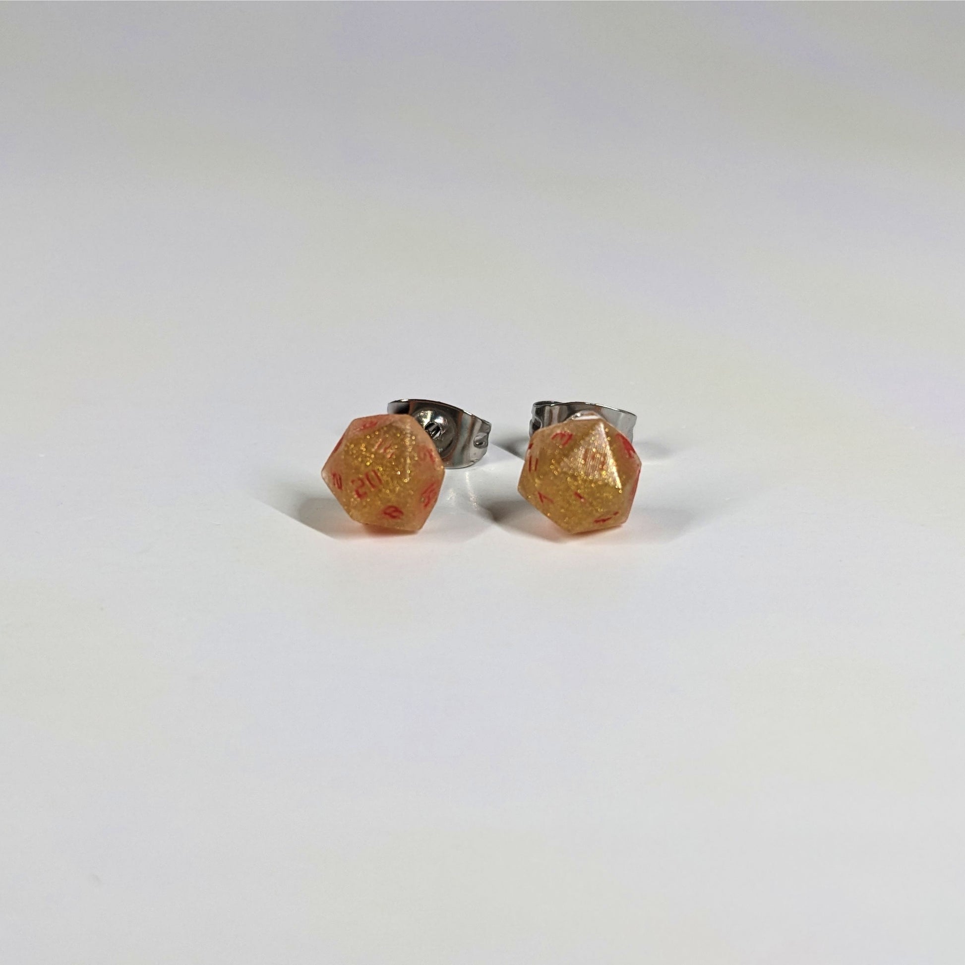 A single pair of the smaller size handmade d20 earrings that are shimmering gold with red numbers on a white background. The earring on the left centers the 20 face and the earring on the right centers the 1 face.