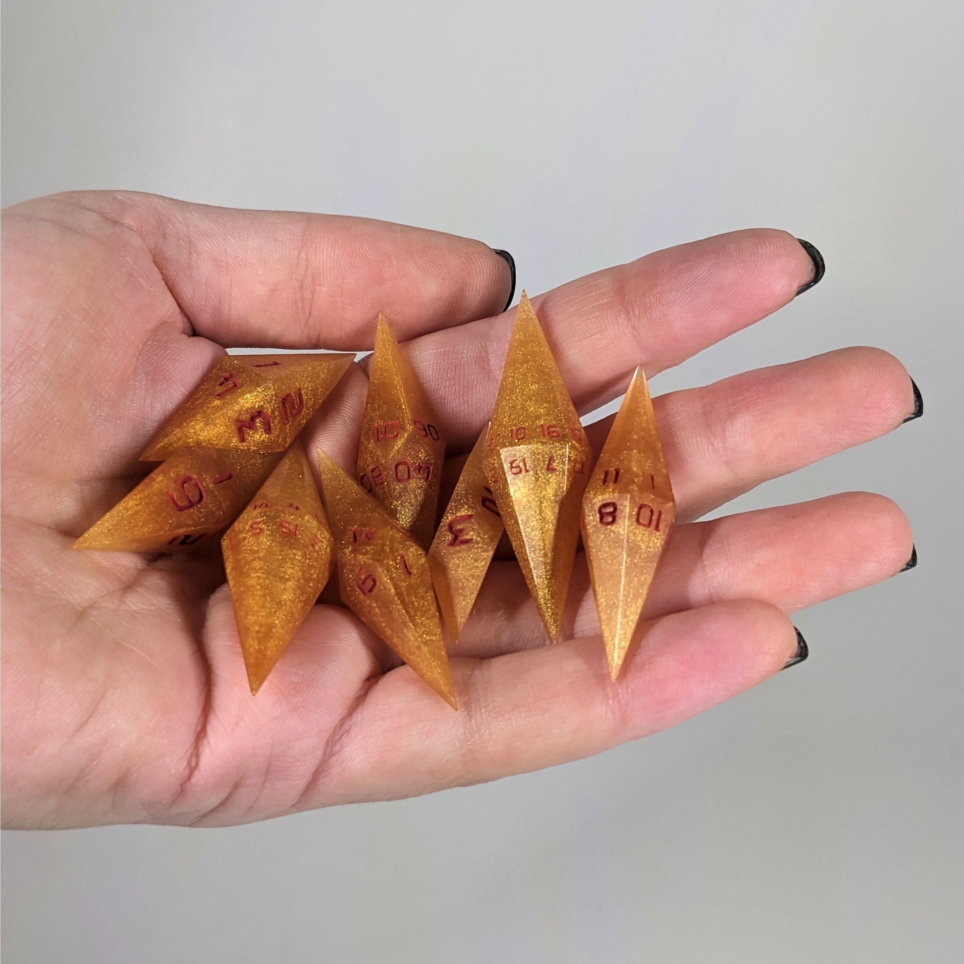 An 8 dice set of diamond style dice that are shimmering gold and red gold swirls and red numbers are held in a hand. The image shows that the set fits easily in a hand, making them overall smaller, though sharper, than a standard set of dnd dice.