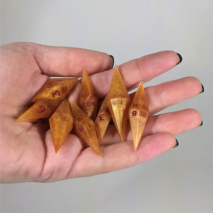 An 8 dice set of diamond style dice that are shimmering gold and red gold swirls and red numbers are held in a hand. The image shows that the set fits easily in a hand, making them overall smaller, though sharper, than a standard set of dnd dice.