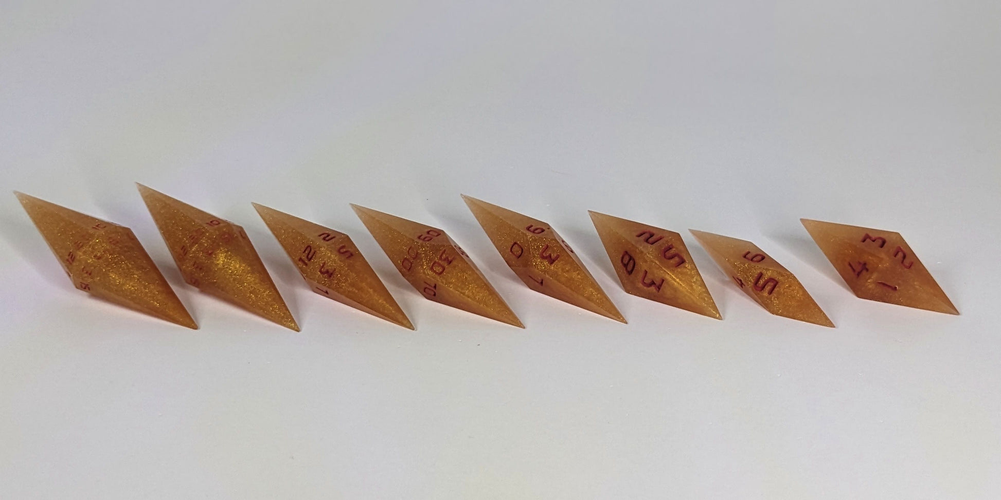 A mid angle photo of a complete set of handmade diamond DnD dice that are shimmering gold and red gold swirls and red numbers arranged in a straight line with bottom points angled slightly forwards and towards the right. They are lined up with the 2 d20s on the left going down to the d4 on the right side. The highest value number of each die is facing forward.