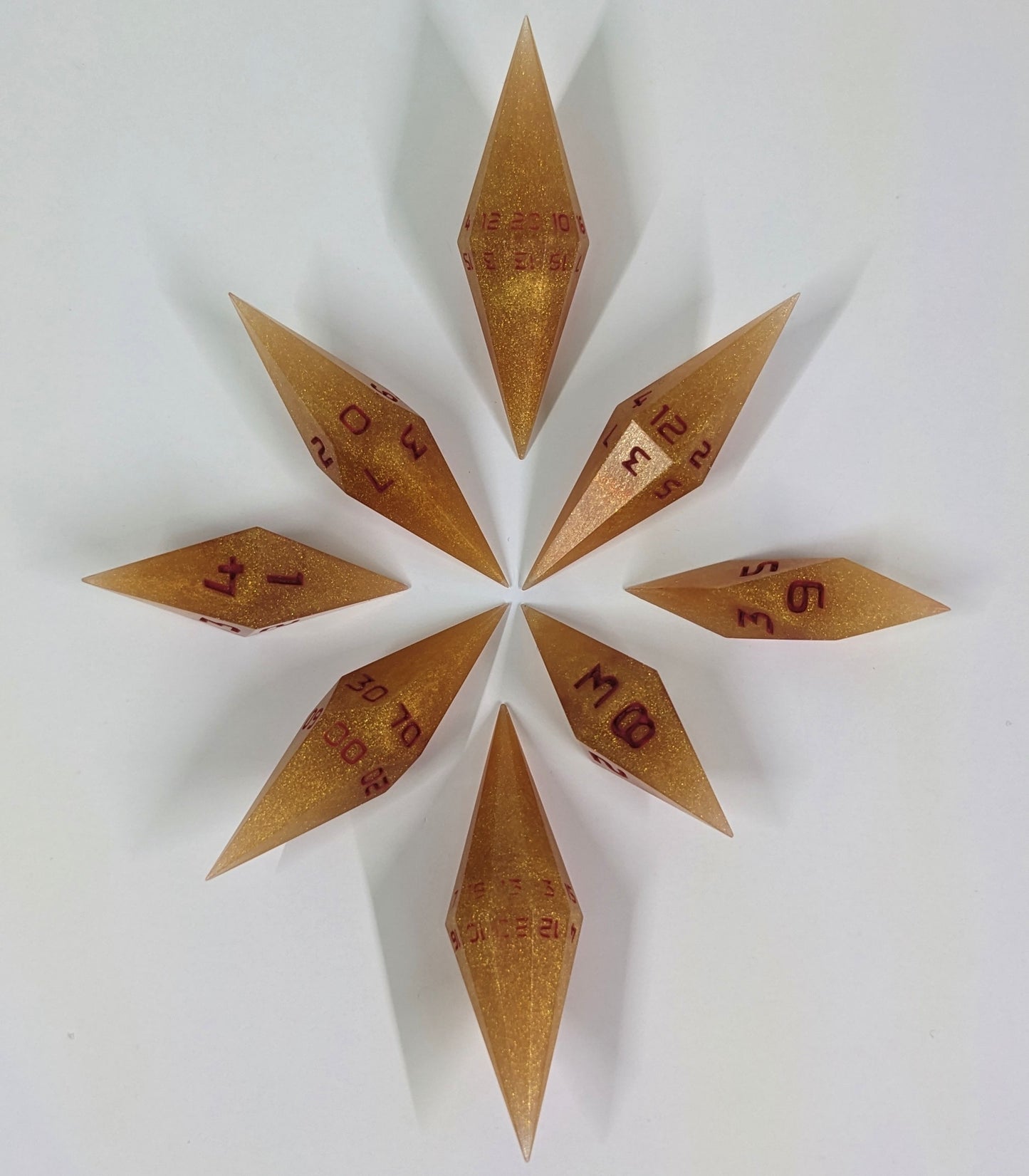 A complete set of diamond shaped resin dice featuring 2 d20s. These handmade dice are shimmering gold and red gold swirls and red numbering and are clustered in a circle with the bottom points angled toward a central point on a white background.