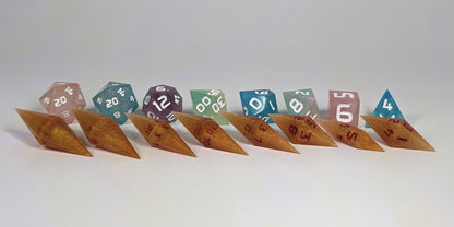 A straight on photo of a complete set of dagger dice that are shimmering gold and red gold swirls and red numbers arranged in a straight line with points angled forward and to the right. They are lined up with the 2 d20s on the left going down to the d4 on the right side. Behind the dagger set is a standard set of polyhedral dice to allow for size comparison. The diamond dice are much smaller overall. The highest value number of each die is facing forward.