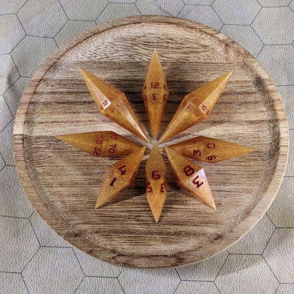 A set of shimmering gold and red gold swirls and red numbering handmade dice arranged on a wooden circle. The resin dice feature a unique pointed diamond shape with the tips facing outward, and the highest number of each die is facing up.