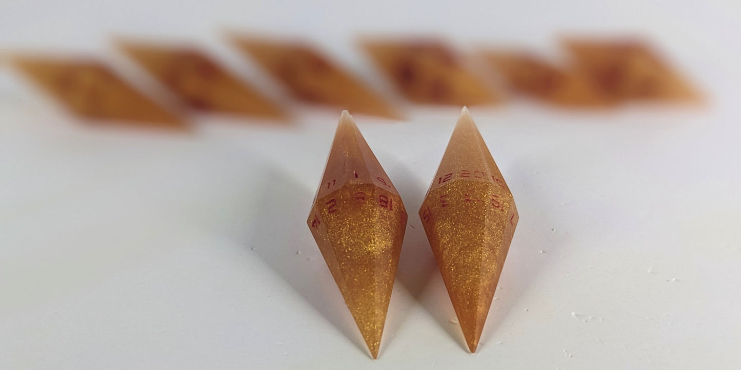 A straight on photo of a diamond set of DnD dice that are shimmering gold and red gold swirls and red numbers focusing on 2 d20s in the center. The d20 on the left has the 1 face up and the d20 on the right has the 20 face up. The other 6 dice are lined up in the background but are out of focus, while the d20s are in sharp focus.