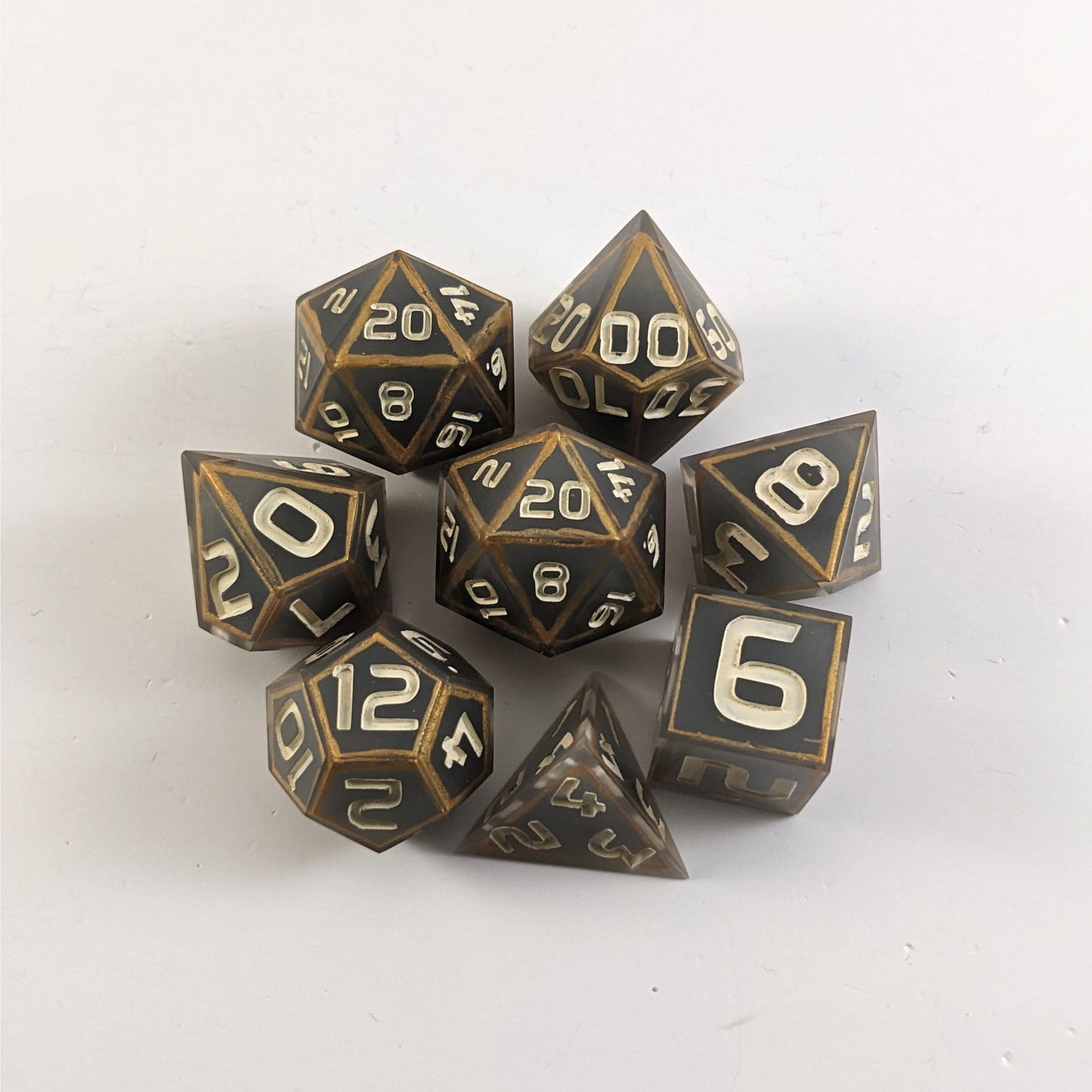 A complete set of 8 handmade artisan dice that are gray with subtle marbling with painted gold lines along the edges a clear shell with ivory numbers. The DnD dice set is arranged in a circle and features 2 D20s, one of which is in the center surrounded by the other 7 dice.