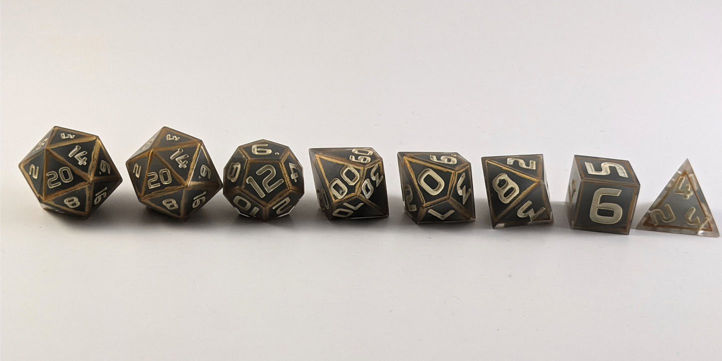 A straight on photo of a complete set of polyhedral dice that are gray with subtle marbling with painted gold lines along the edges a clear shell with ivory numbers arranged in a straight line. They are lined up with the 2 d20s on the left going down to the d4 on the right side. The highest value number of each die is facing forward.