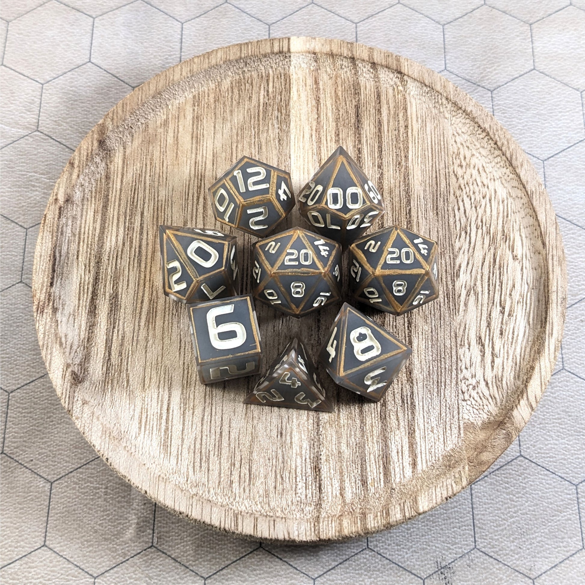 A complete set of 8 handmade artisan dice that are gray with subtle marbling with painted gold lines along the edges in a clear shell with ivory numbers. The DnD dice set is on a wooden circle and the dice are arranged in a circle and features 2 D20s, one of which is in the center surrounded by the other 7 dice.