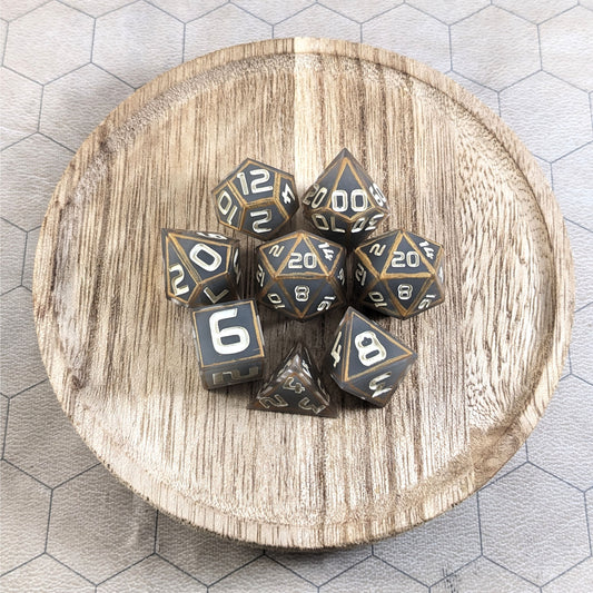 A complete set of 8 handmade artisan dice that are gray with subtle marbling with painted gold lines along the edges in a clear shell with ivory numbers. The DnD dice set is on a wooden circle and the dice are arranged in a circle and features 2 D20s, one of which is in the center surrounded by the other 7 dice.
