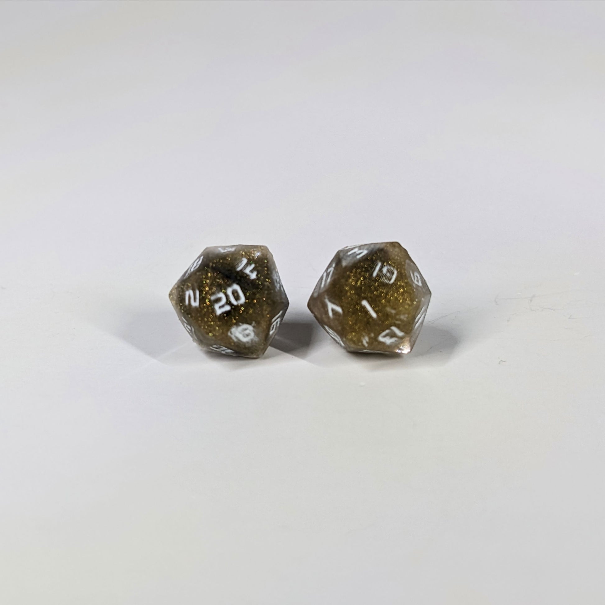 A single pair of the larger size handmade d20 earrings with sterling silver hardware that are glittering gold and black with white numbers on a white background. The earring on the left centers the 20 face and the earring on the right centers the 1 face.
