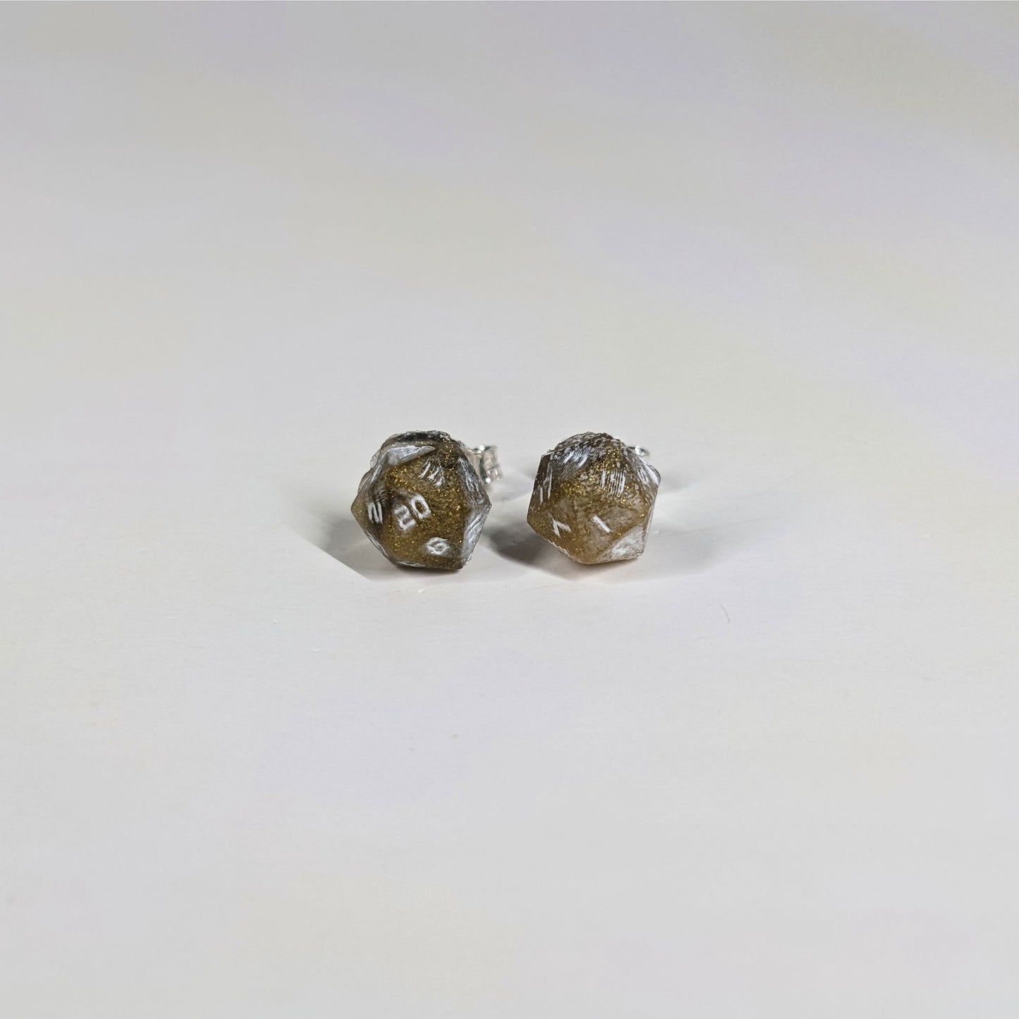 A single pair of the smaller size handmade d20 earrings with sterling silver hardware that are glittering gold and black with white numbers on a white background. The earring on the left centers the 20 face and the earring on the right centers the 1 face.