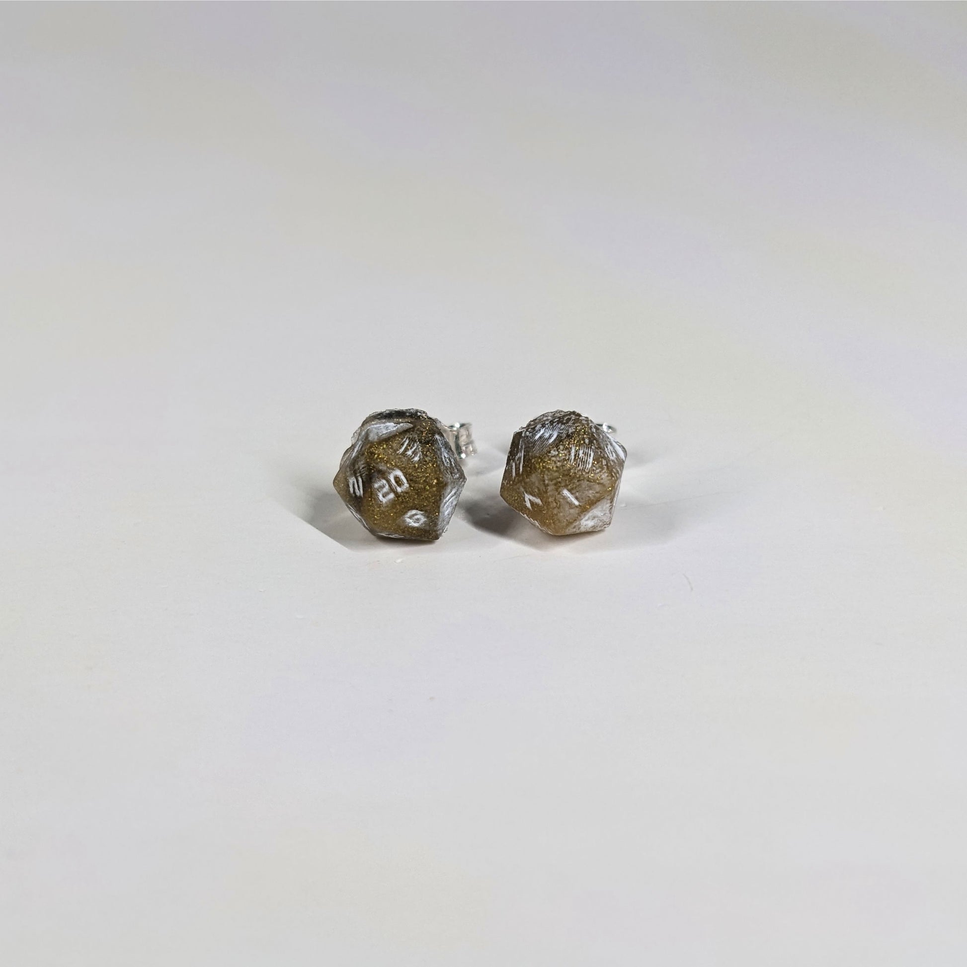 A single pair of the smaller size handmade d20 earrings with sterling silver hardware that are glittering gold and black with white numbers on a white background. The earring on the left centers the 20 face and the earring on the right centers the 1 face.