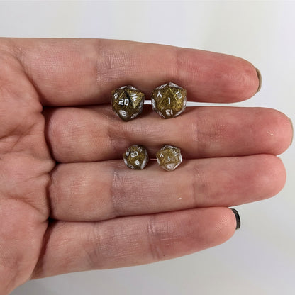 Two pairs of handmade d20 earrings with sterling silver hardware that are glittering gold and black with white numbers held between the fingers of a flat hand. The larger pair is between the pointer and middle finger, the smaller between the middle and ring finger. The earrings on the left of each pair centers the 20 face and the earrings on the right of each pair centers the 1 face.