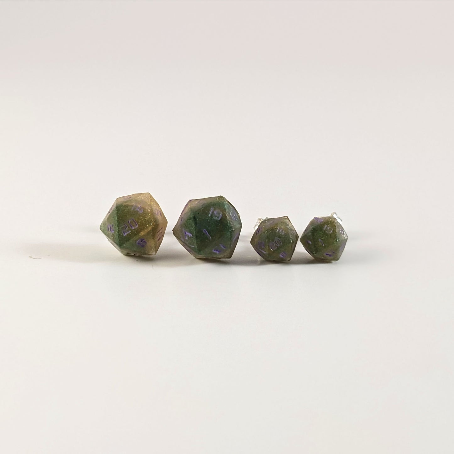 Two pairs of handmade d20 earrings with sterling silver hardware that are dark green and bright gold with purple numbers on a white background sit in a line with the larger pair on the left and the smaller pair on the right. The earrings on the left of each pair centers the 20 face and the earrings on the right of each pair centers the 1 face.