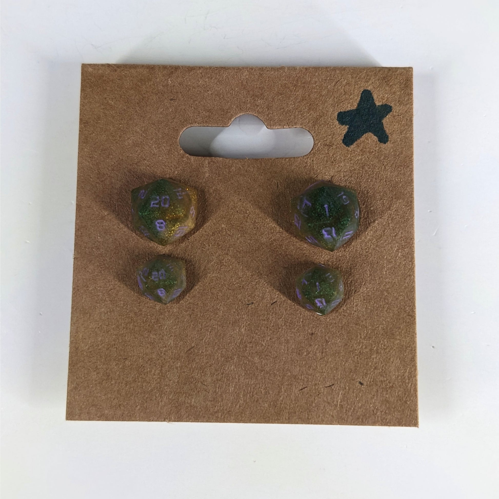 Two pairs of handmade d20 earrings with sterling silver hardware that are dark green and bright gold with purple numbers on a kraft brown earring card with a star in the top right corner. The earrings on the left of each pair centers the 20 face and the earrings on the right of each pair centers the 1 face.