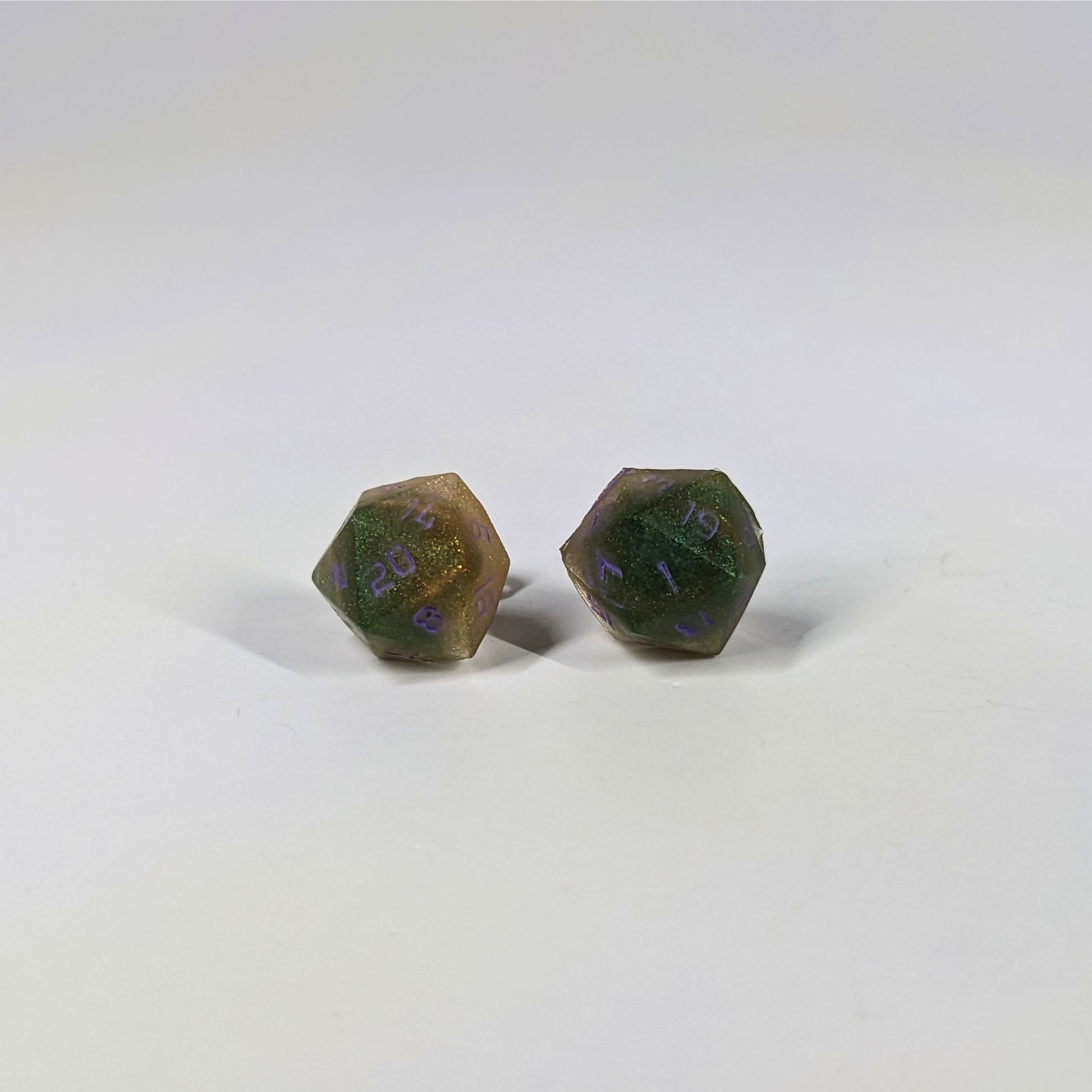 A single pair of the larger size handmade d20 earrings with sterling silver hardware that are dark green and bright gold with purple numbers on a white background. The earring on the left centers the 20 face and the earring on the right centers the 1 face.