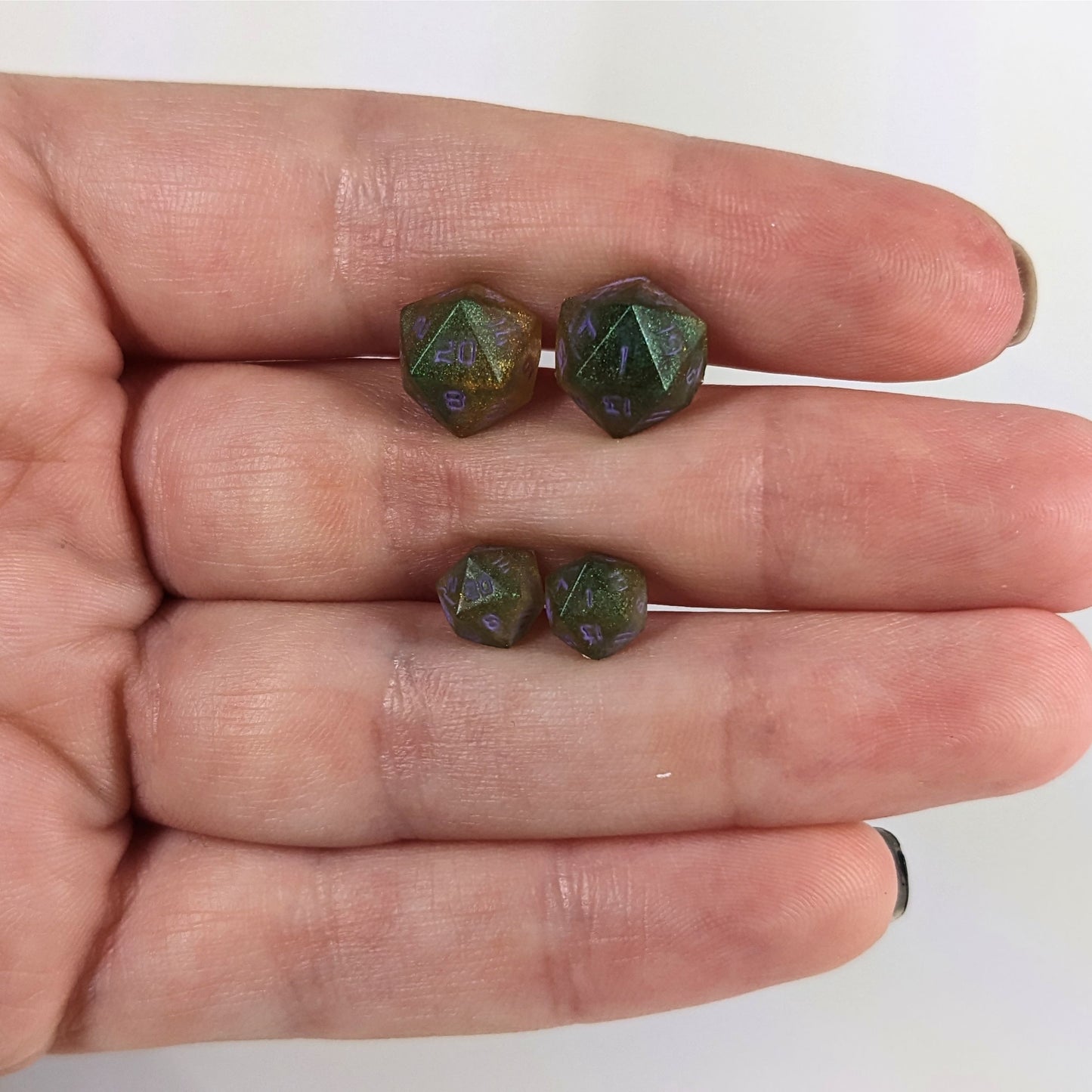 Two pairs of handmade d20 earrings with sterling silver hardware that are dark green and bright gold with purple numbers held between the fingers of a flat hand. The larger pair is between the pointer and middle finger, the smaller between the middle and ring finger. The earrings on the left of each pair centers the 20 face and the earrings on the right of each pair centers the 1 face.