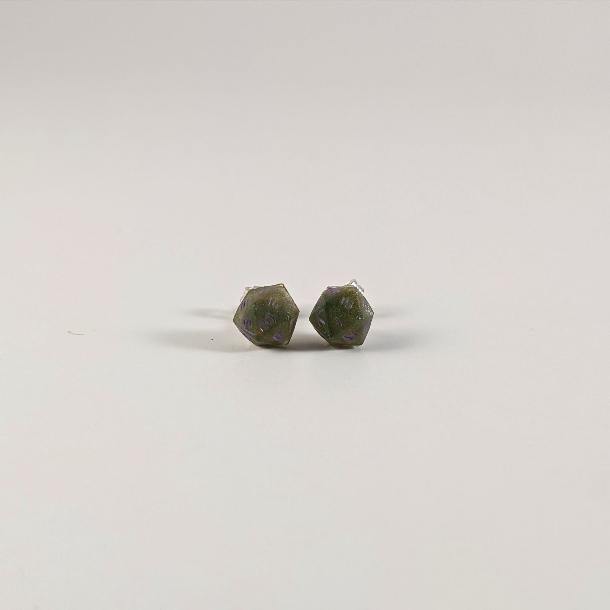 A single pair of the smaller size handmade d20 earrings with sterling silver hardware that are dark green and bright gold with purple numbers on a white background. The earring on the left centers the 20 face and the earring on the right centers the 1 face.