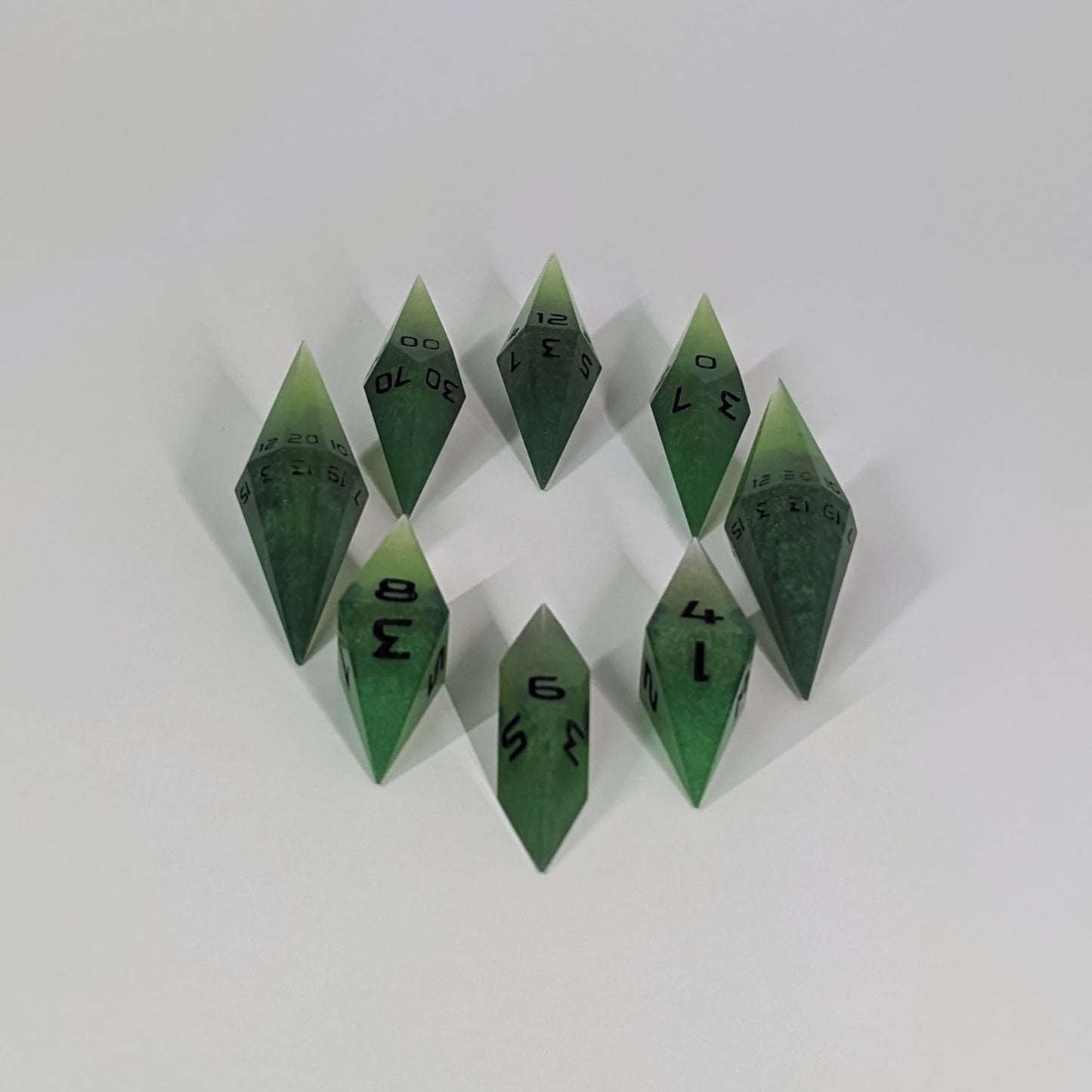 A complete set of resin dice featuring 2 d20s for advantage rolls. These swirling shades of green with black numbering slender handmade diamond dice are clustered in a tight circle on a white background, highlighting the quality of the artisan dice craftsmanship and the unique pointed shape.
