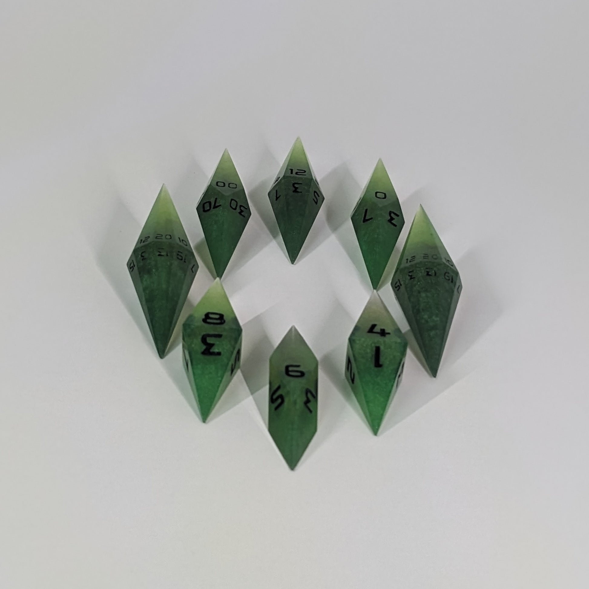 A complete set of resin dice featuring 2 d20s for advantage rolls. These swirling shades of green with black numbering slender handmade diamond dice are clustered in a tight circle on a white background, highlighting the quality of the artisan dice craftsmanship and the unique pointed shape.