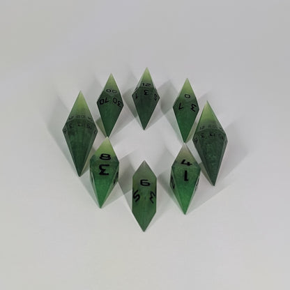 A complete set of resin dice featuring 2 d20s for advantage rolls. These swirling shades of green with black numbering slender handmade diamond dice are clustered in a tight circle on a white background, highlighting the quality of the artisan dice craftsmanship and the unique pointed shape.