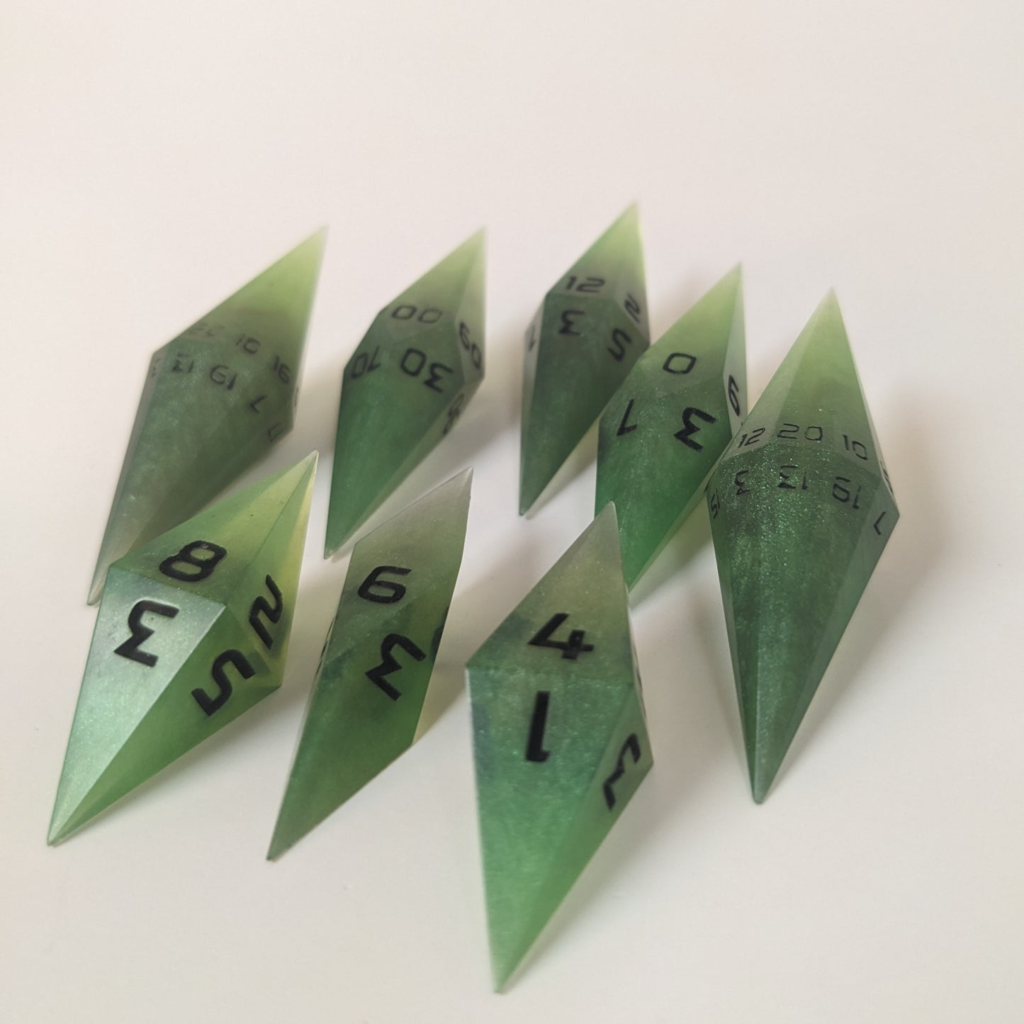 A complete set of resin dice featuring 2 d20s for advantage rolls. These swirling shades of green with black numbering slender handmade diamond dice are clustered in a tight circle on a white background, highlighting the quality of the artisan dice craftsmanship and the unique pointed shape.