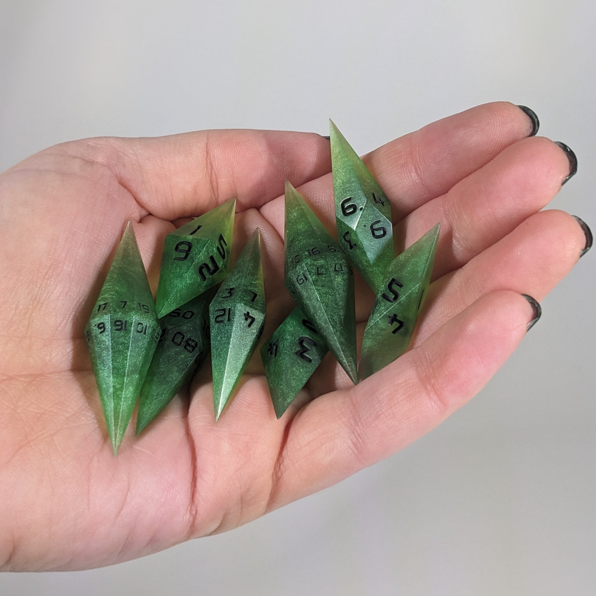 Close-up of swirling shades of green with black numbering artisan dice resting in a palm on a seamless white backdrop. The handmade dice feature a distinct sharp diamond shape, with 2 d20s visible among the high-quality resin dice.