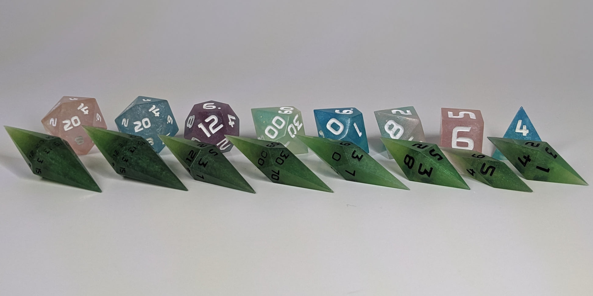A size comparison of swirling shades of green with black numbering long pointed diamond handmade dice with standard shapes placed behind for scale. The photo emphasizes the length of the diamond dice being much greater than a standard set as well as the diamond set being much more slender. This artisan dice set features 2 d20s and rests on a seamless white background to highlight the elongated profile of the resin dice.